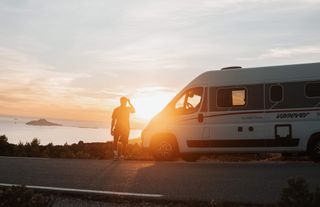 Ein Mann geht zum Camper, im Hintergrund ein Sonnenuntergang über dem Meer